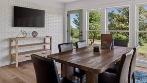 Un comedor con una mesa de madera, sillas negras, un televisor montado en una pared de ladrillo blanco y vistas a zonas verdes al exterior.