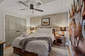 A bedroom with a bed, two bedside tables with lamps, a ceiling fan, a door, and a wall with a painting.