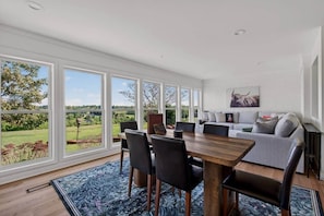 A spacious room with a dining table, chairs, and a sofa. Large windows offer a view of the outdoors.