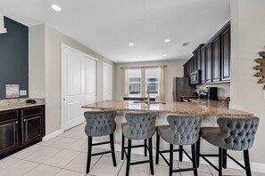 A kitchen with a central island, bar stools, dark cabinets, and a view to the outside. - Themed Home with Game Room and Private Pool near Disney with Waterpark Access. (Kissimmee)