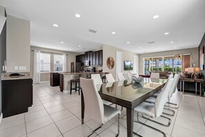 A modern kitchen with dark cabinets, a dining area with a glass table and white chairs, and a living area with a sofa and lamp. - Themed Home with Game Room and Private Pool near Disney with Waterpark Access. (Kissimmee)