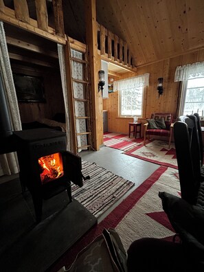 A cozy cabin interior with a wood-burning stove, a dining area, and a window with a view of trees.