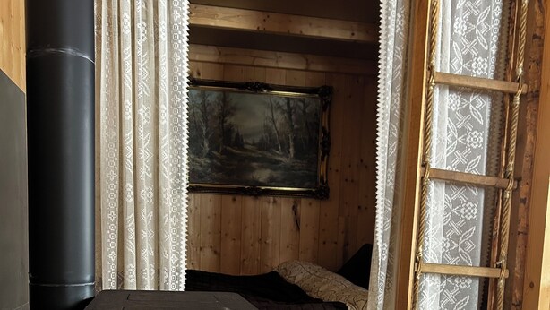 Una estufa de leña con llamas visibles, una cama con una colcha oscura, un muro de madera y una cortina estampada.