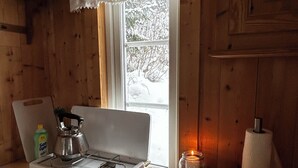Interior de una cabaña de madera con estufa, ventana con cortina de encaje, vela y dispensador de toallas de papel.