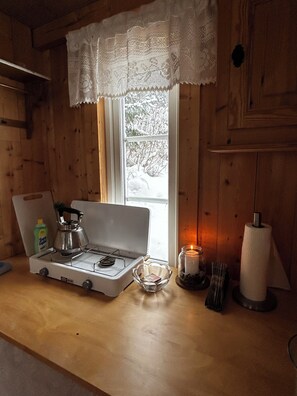 A wooden cabin interior with a stove, a window with a lace curtain, a candle, and a paper towel holder.