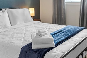 Un lit bien fait, avec du linge de lit blanc et une couverture bleue, une pile de serviettes blanches et une table de chevet avec une lampe.
