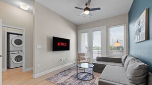 A modern living room with a flat-screen TV, a ceiling fan, a sofa, a coffee table, and a view through large windows.