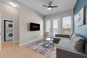 Una sala de estar moderna con una televisión de pantalla plana, un ventilador de techo, un sofá, una mesa de centro y vistas a través de ventanas amplias.
