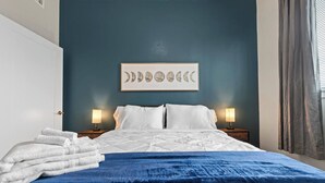 A neatly made bed with white linens, a blue throw blanket, and a wall art piece depicting moon phases.