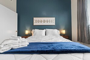 Un lit bien fait, avec du linge de lit blanc, un plaid bleu et une œuvre d’art murale représentant les différentes phases de la lune.