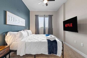 Une chambre à coucher avec un lit, un ventilateur de plafond, une télévision et une fenêtre avec des rideaux.