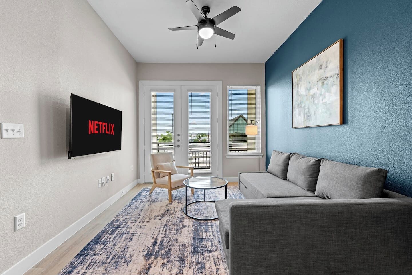 A living room with a flat-screen TV, a ceiling fan, a sofa, a chair, a coffee table, and a balcony with windows.