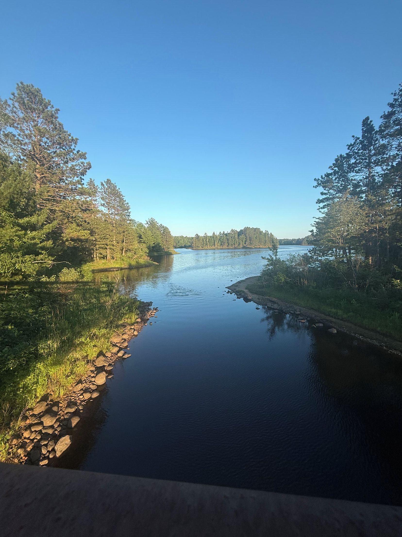 Een kalme rivier met bomen aan beide oevers, een heldere lucht en een rotsachtige oever.