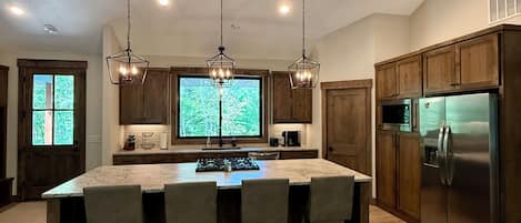 A modern kitchen with a central island, pendant lighting, wooden cabinets, and a stainless steel refrigerator.