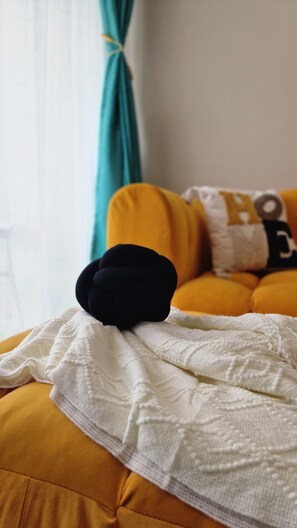 Ein Bett mit weißer Decke, einem schwarzen Kopfkissen, einem gelben Kissen und einem blaugrünen Vorhang in einem Zimmer mit weißer Wand.