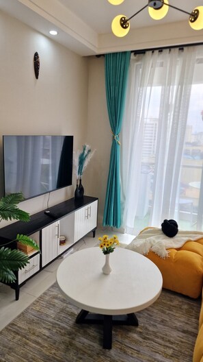 Ein modernes Wohnzimmer mit einem Flachbildfernseher, einem runden weißen Tisch mit Blumen in einer Vase, einem gelben Sofa und türkisblauen Vorhängen.