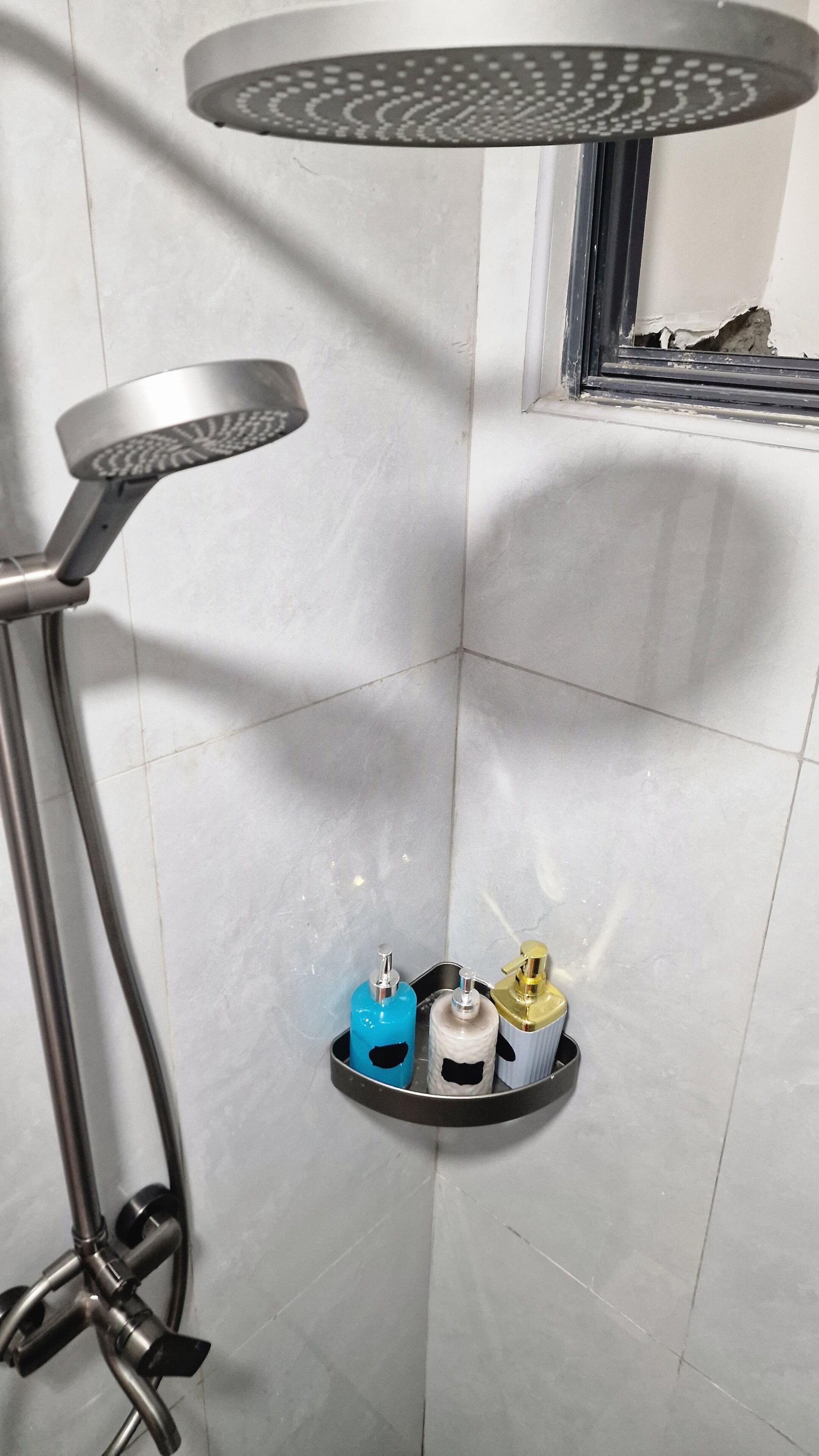 Zona doccia con soffione a mano, una finestra con un vetro rotto e uno scaffale con tre flaconi di articoli da toilette.