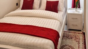 A neatly made bed with white and red bedding, a bedside table with a vase of flowers, and a patterned rug. - Stylish High-Rise Retreat, Skyline Views, Gym & Rooftop in Kilimani (Nairobi)