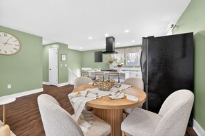 Een eetruimte met een ronde tafel, stoelen, een klok en een keuken op de achtergrond.