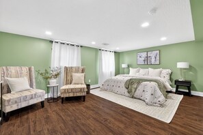 Een slaapkamer met een bed, twee stoelen, een nachtkastje, een lamp en een kunstwerk aan de muur.