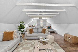 Ein Loft-Wohnzimmer mit hohen Decken, einem grauen Sofa mit braunen Kissen, einem Couchtisch mit einer Zeitschrift, einer Pflanze und einem Korbwäschesammler.