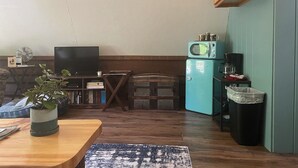 A compact living space with a wooden dining table, a television, a small kitchen area with a microwave, and a trash bin. - A-Frame Cabin -The Get Away #1 (Diana)