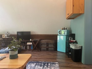 Un espacio habitable compacto con una mesa de comedor de madera, un televisor, una pequeña cocina con microondas y un cubo de basura.