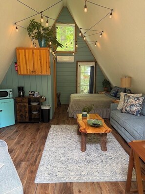 A cozy attic bedroom with a sloped ceiling, a bed, a wooden cabinet, a small kitchenette, and string lights. - A-Frame Cabin -The Get Away #1 (Diana)