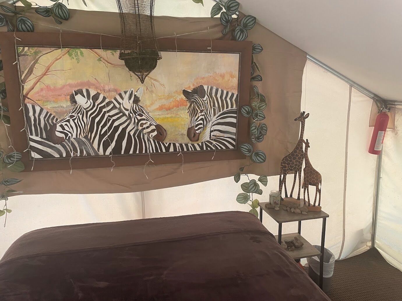 A tent with a bed, a painting of zebras, a giraffe sculpture, and hanging plants.