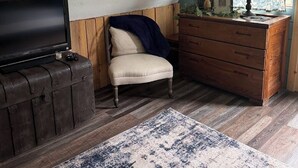 Ein gemütliches Zimmer mit Fernseher auf einer Holzkiste, einem Sessel mit Kissen, einem hölzernen Schminktisch und einem gemusterten Teppich.