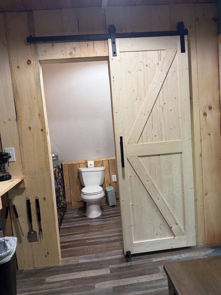 A rustic bathroom with a wooden door, a toilet, and a small shelf with tools.