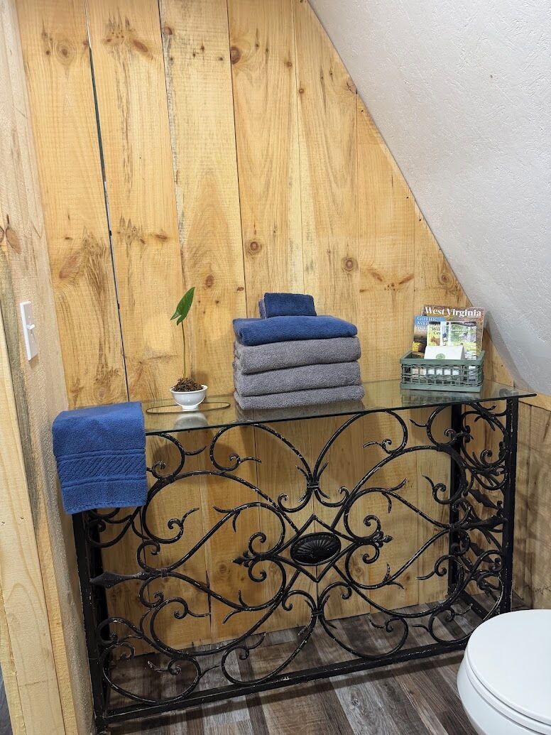 A bathroom with a wooden sloped ceiling, a glass-top vanity with a black wrought iron frame, a white toilet, and a magazine on the vanity.