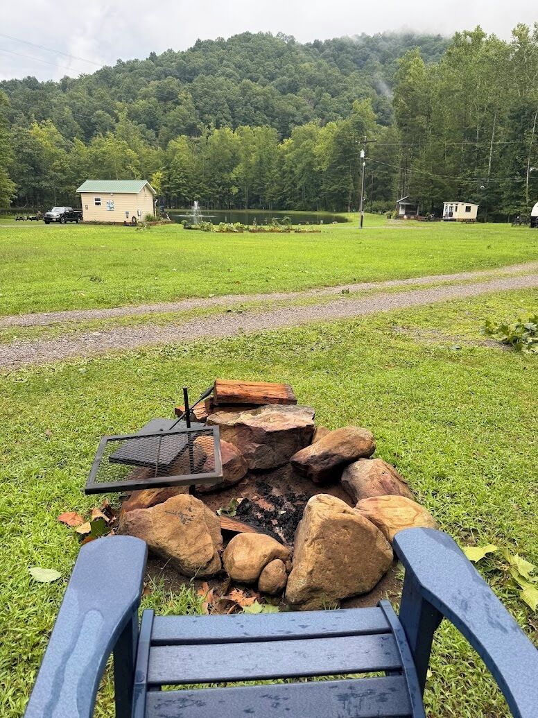 A campsite with a fire pit surrounded by rocks, a bench, and a view of a forested area with cabins in the background.