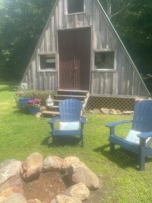 A wooden cabin with a front door, two Adirondack chairs, a fire pit, and a wooden fence.