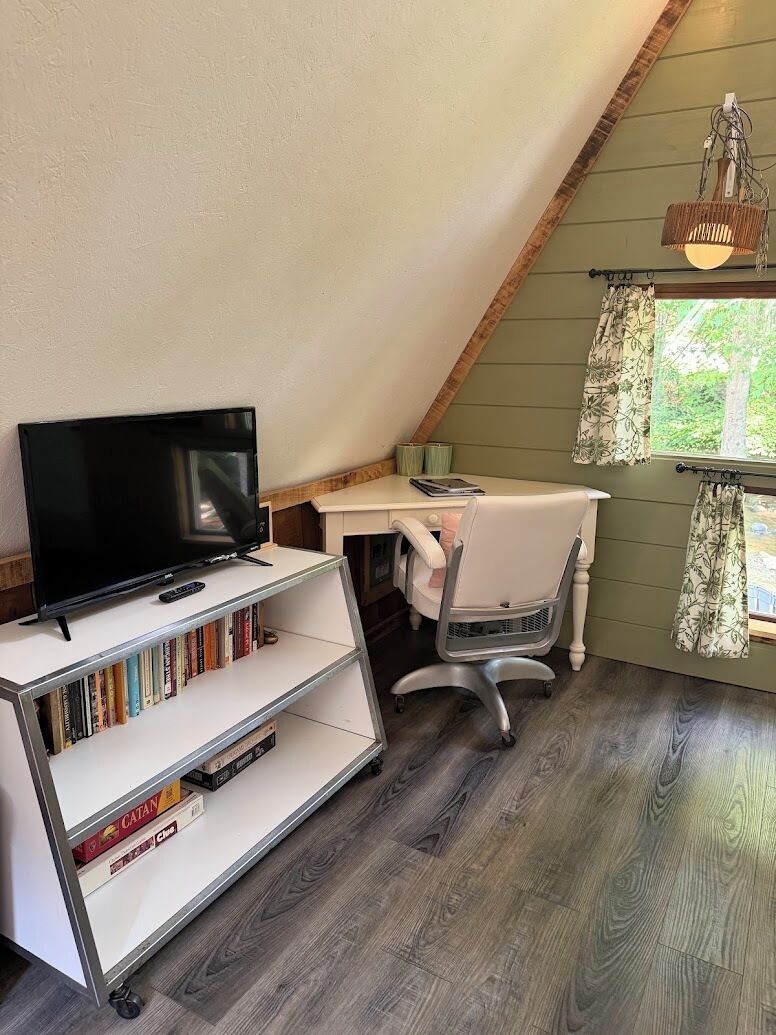 Una habitación con techo inclinado, un televisor sobre un soporte blanco, un escritorio con una silla y una ventana con cortinas.