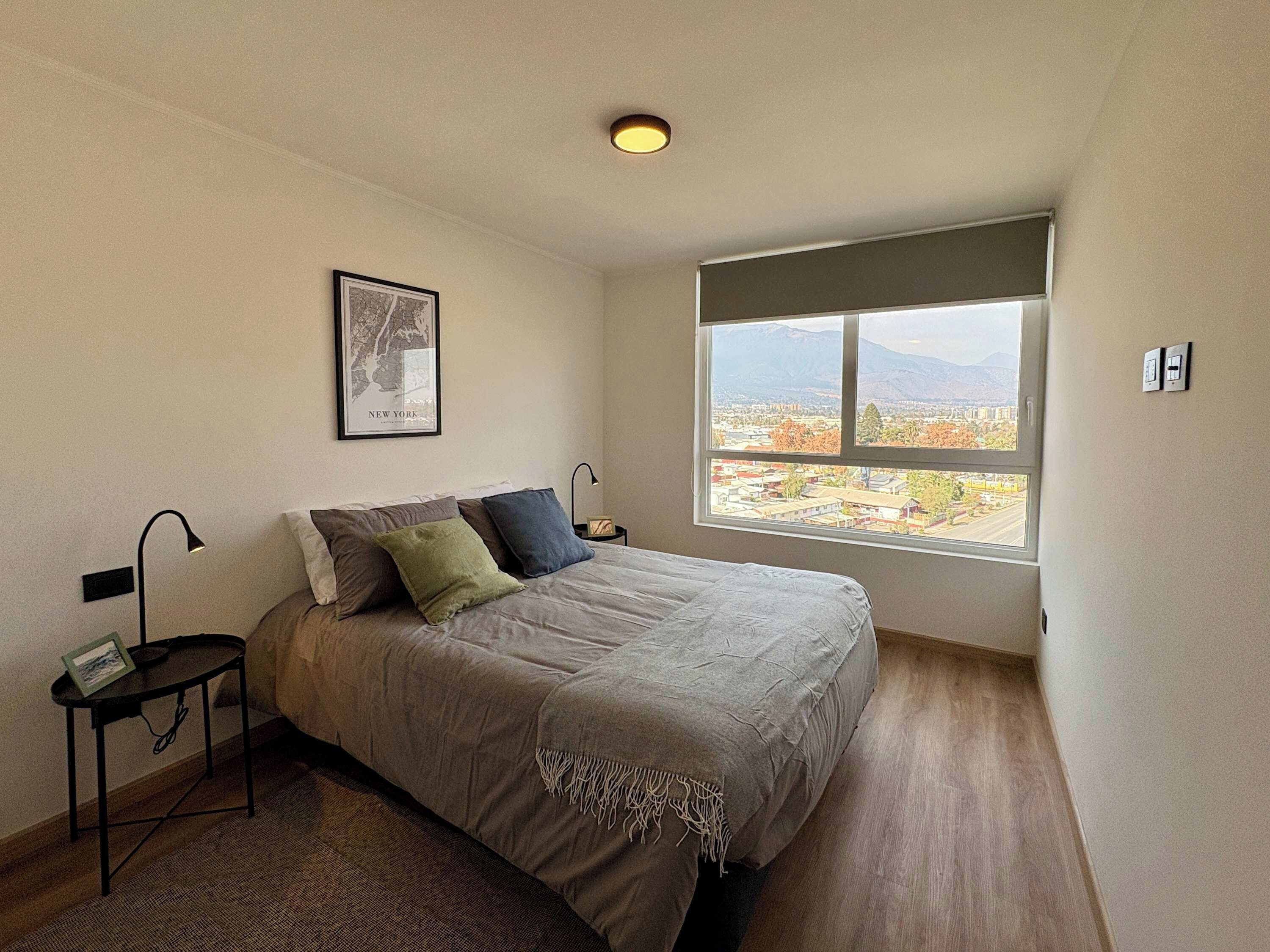 Un dormitorio con cama, mesita de noche, lámpara, un cuadro en la pared y una ventana con vistas a un paisaje urbano y montañas.