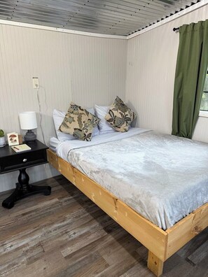 A single bed with a wooden frame and a grey mattress, a bedside table with a lamp, and a power outlet on the wall.