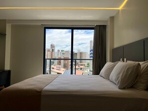 A hotel room with a large bed, a view of the cityscape through a large window, and a modern design. - Vert House - Apartment near the beach in CariBessa for 4 people (João Pessoa)