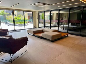 A modern hotel lobby with a sectional sofa, a small table, and a large window offering a view of the outdoor area. - Vert House - Apartment near the beach in CariBessa for 4 people (João Pessoa)