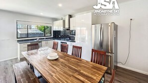 A modern kitchen with white cabinets, a wooden dining table, and a stainless steel refrigerator. - Jerrara Escape Two - Great for the whole Family (East Jindabyne)
