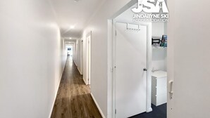A hotel corridor with white walls, wooden flooring, and a closed door with a small shelf. - Jerrara Escape Two - Great for the whole Family (East Jindabyne)
