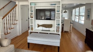A living room with a large flat-screen TV, wooden floors, a staircase, and a dining area.