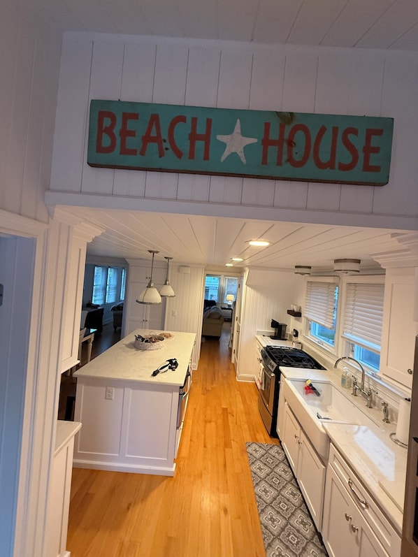 Cocina con isla central, muebles blancos, estufa y pileta. Un cartel indica 'BEACH HOUSE'.