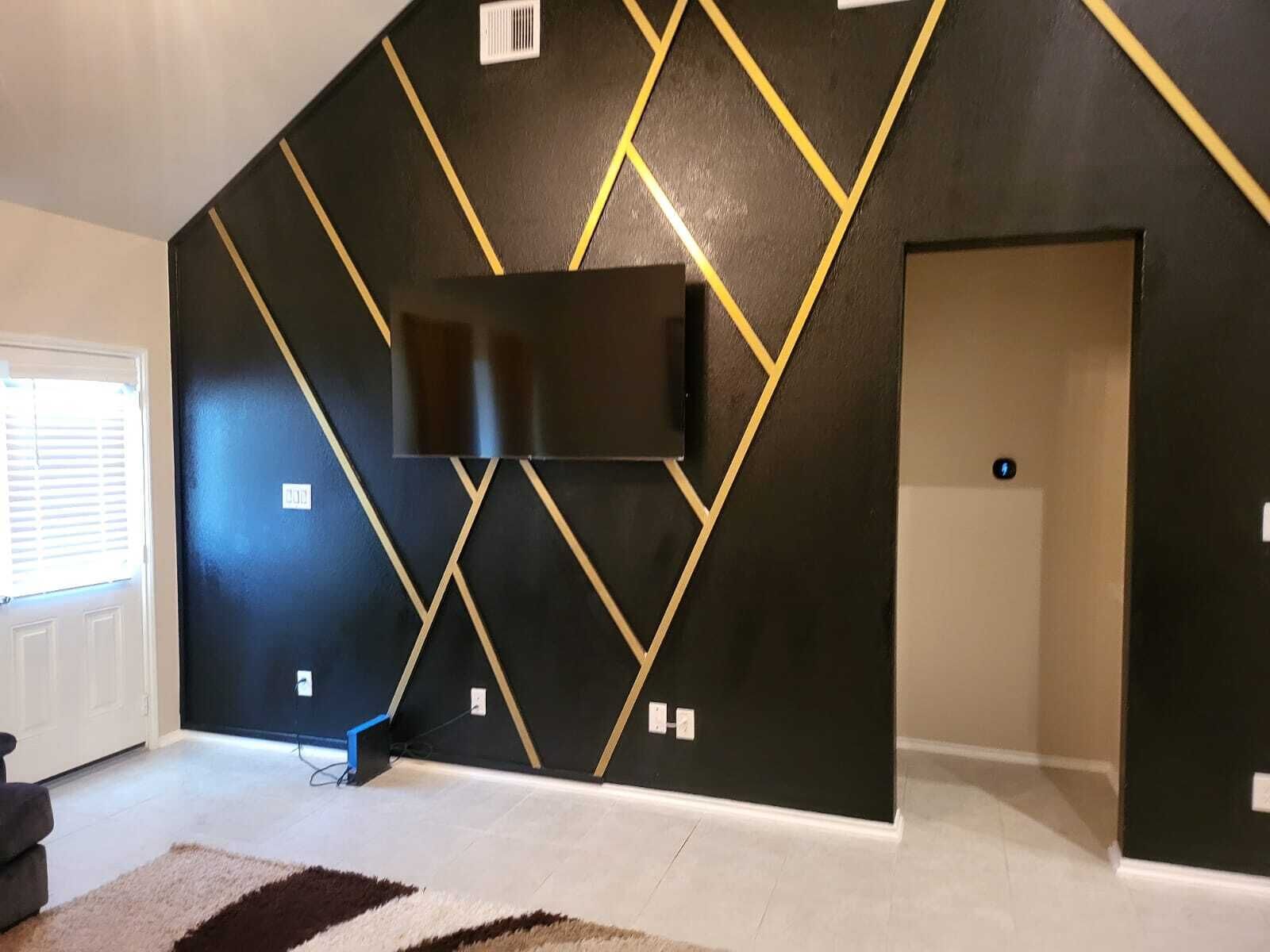 A modern living room with a black and gold geometric wall design, a flat-screen TV mounted on the wall, and a door to the right.