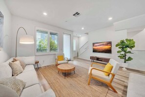 Ein modernes Wohnzimmer mit einer Couch, Sesseln, einem runden Couchtisch, einer Stehlampe, einem Fernseher und einer Treppe.