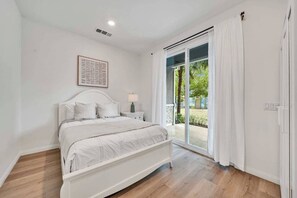 Ein Schlafzimmer mit einem Bett, einem Nachttisch, einer Lampe, einem gerahmten Bild und einer Schiebetür, die auf eine Terrasse führt.