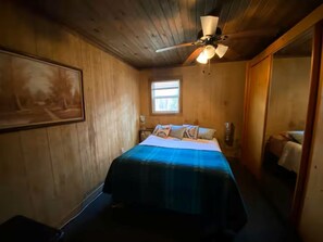 A bedroom with wooden walls, a bed with a blue bedspread, a ceiling fan, and a window. - #12 Nice pondside cabin on the river (Diana)