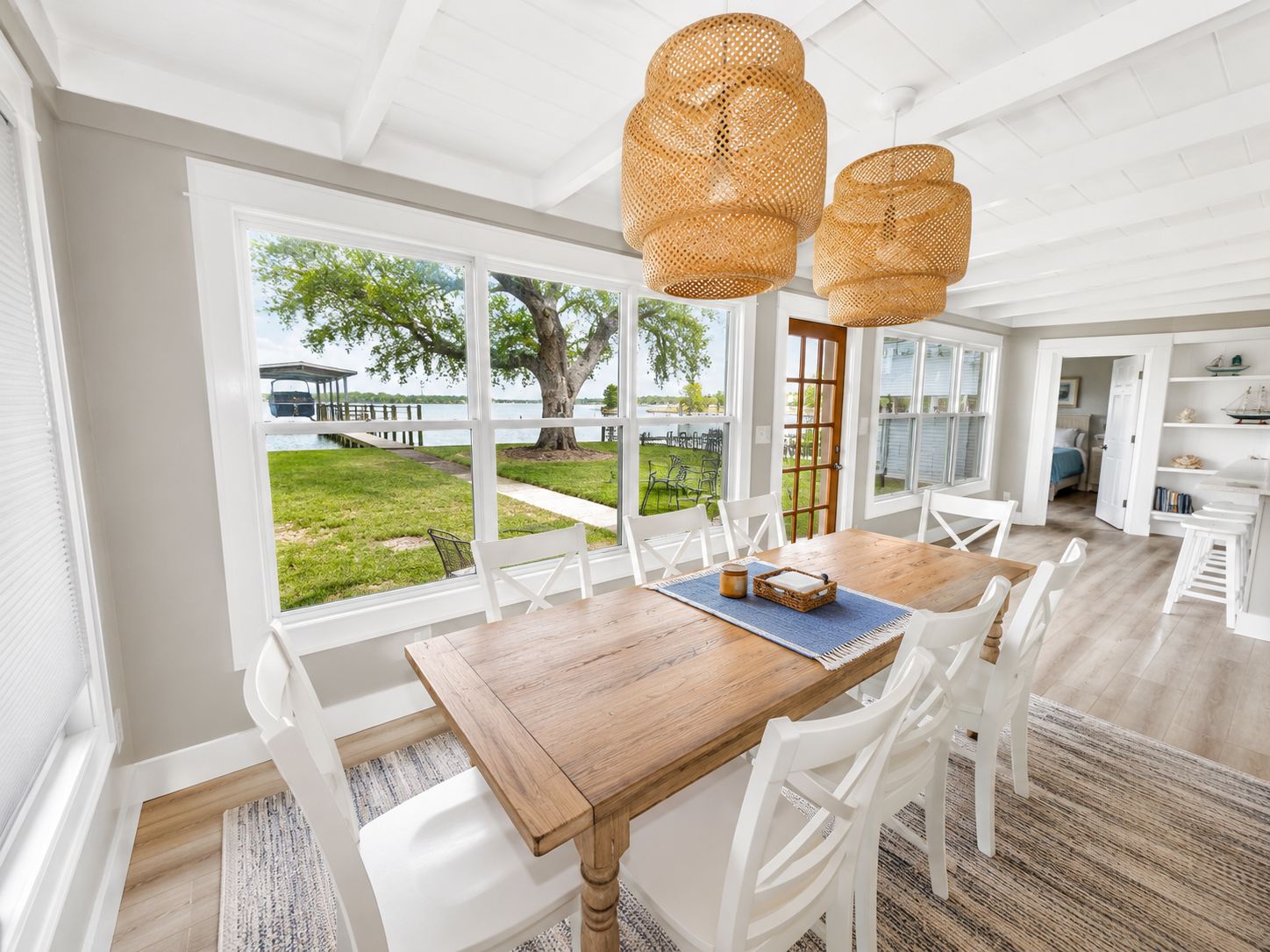 A dining area with a wooden table and white chairs, large windows with a view of a tree and a dock, and a ceiling with woven light fixtures.