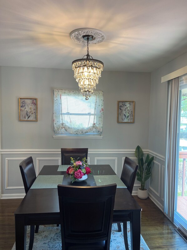 A dining room with a chandelier, floral wall art, a window with blinds, and a potted plant.