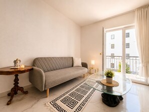 Ein modernes Wohnzimmer mit einer Couch, einem Glastisch mit Pflanze, einem gemusterten Teppich und einem kleinen Tisch mit Kerze und Figur.